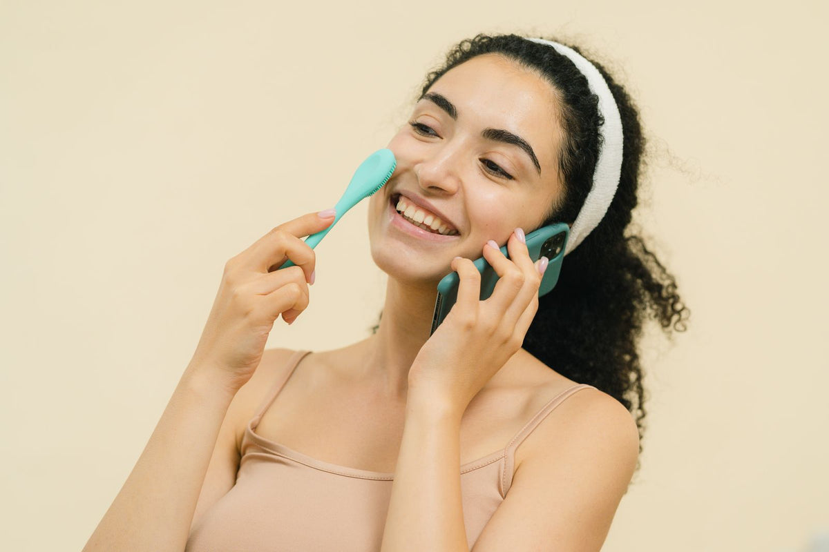 Mujer alegre usando herramienta de skincare mientras habla por celular